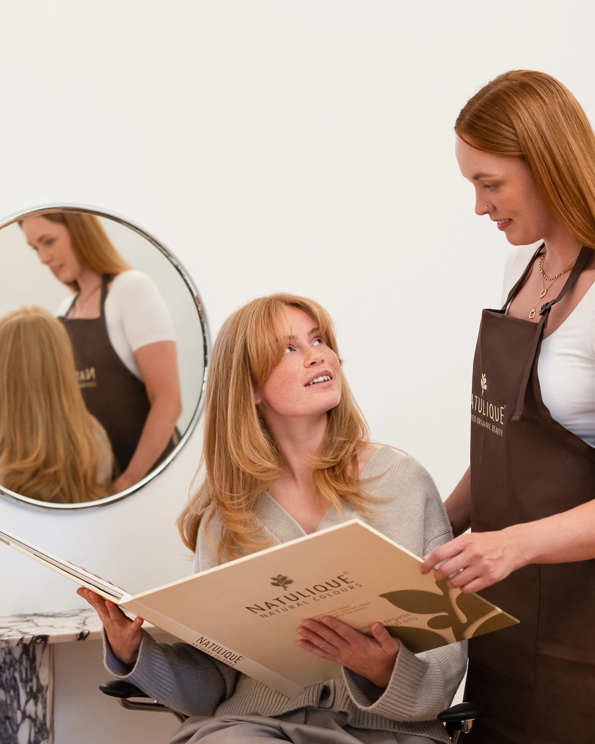 Haas & Winkler Friseure - Eine Friseurin, die eine braune Schürze der Marke NATULIQUE trägt, zeigt einer Kundin Haarfarbenproben aus einem großen Farbmusterheft.